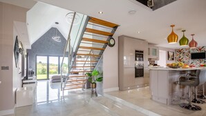 A modern kitchen with a central island, a staircase with wooden steps, and a dining area with bar stools. - 3 Bed in Bryngwran (oc-y44816) (Bodedern)