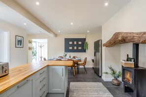 A modern kitchen with a wooden countertop, grey cabinets, a dining area with a table and chairs, and a wood-burning stove. - 4 Bed in Weare Giffard (oc-w44710) (Weare Giffard)