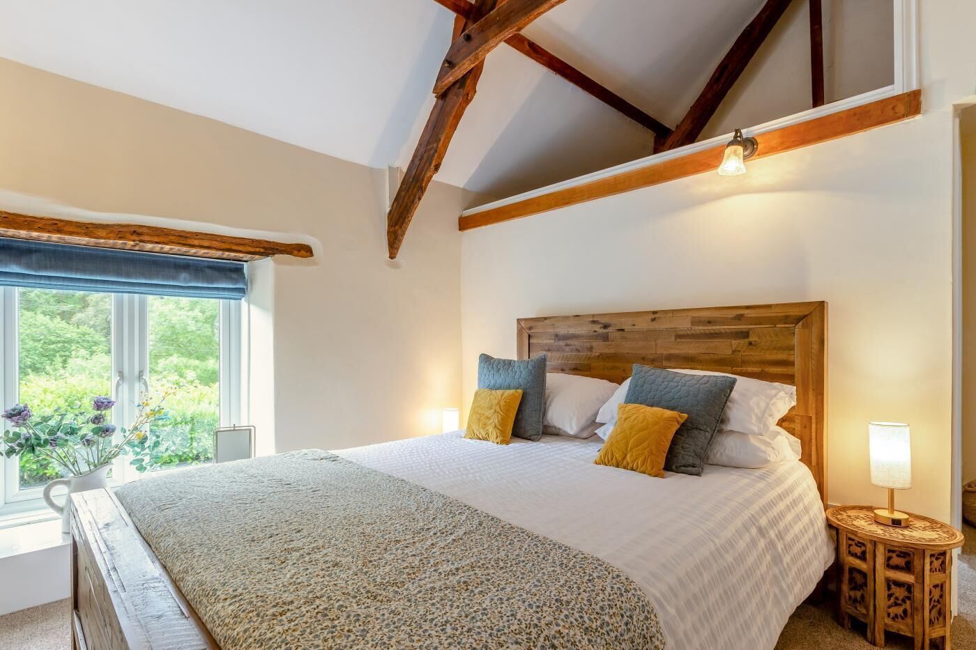 A bedroom with a wooden headboard, a large bed with multiple pillows, a bedside table with a lamp, and a window with a view of greenery.