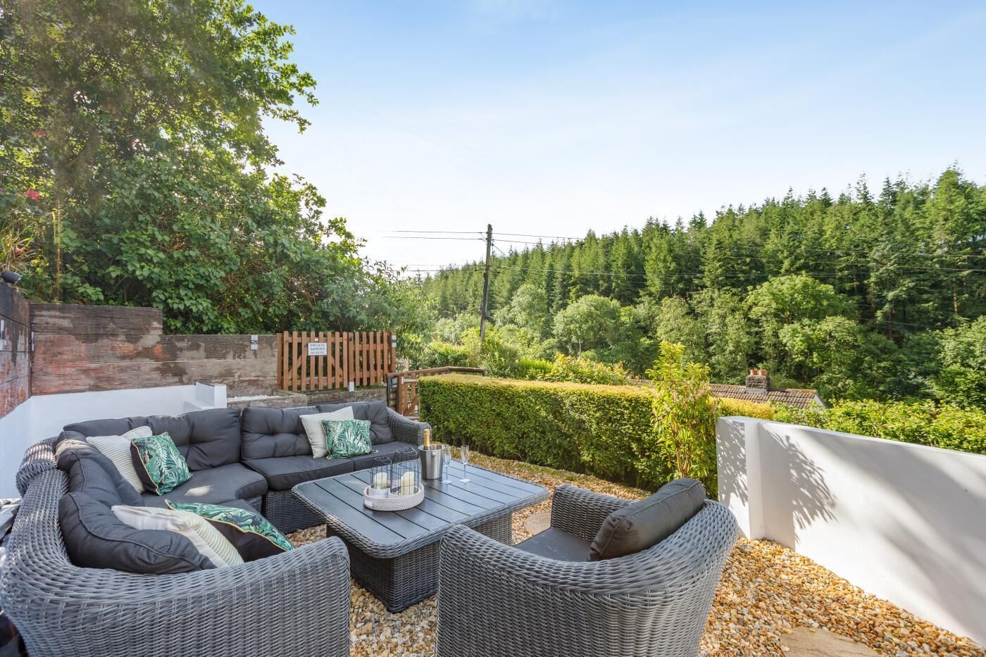 An outdoor seating area with wicker furniture and a table, surrounded by a garden and a wooden fence.