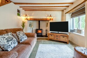 A cozy living room with a brown leather sofa, a flat-screen TV, a wooden entertainment unit, and a stone fireplace. - 4 Bed in Weare Giffard (oc-w44710) (Weare Giffard)