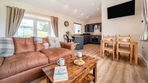 A cozy living room with a brown leather sofa, a wooden coffee table with a book and a cup, and a dining area with a wooden table and chairs. - 2 Bed in Kirkmahoe (oc-h44227) (Dumfries)