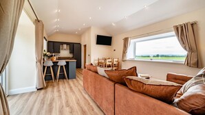 A modern living room with a sofa, a dining area with chairs, a kitchen with a bar, and a large window with a view. - 2 Bed in Kirkmahoe (oc-h44227) (Dumfries)