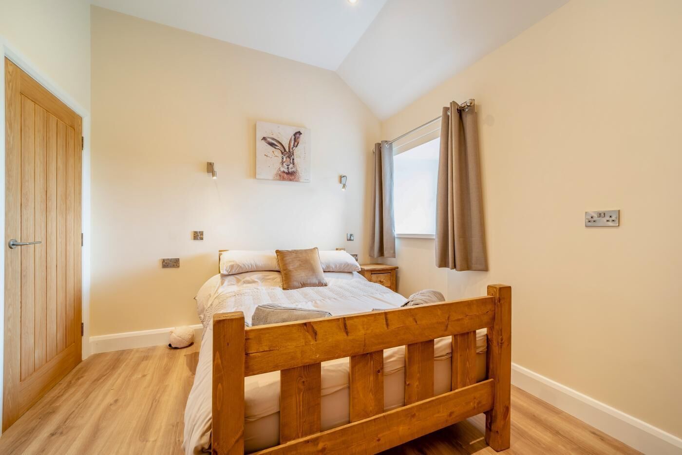 Une chambre à coucher avec un lit en bois, une fenêtre avec des rideaux, une œuvre d’art accrochée au mur et une porte en bois.