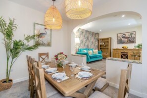 A dining area with a wooden table set for six, surrounded by chairs. There is a large potted plant to the left and a teal sofa in the background. - 5 Bed in Pocklington (oc-m45222) (Pocklington)