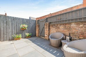 Ein Patio mit Ziegelmauer, Holzlattenzaun und Gartenmöbeln, darunter Korbstühle und ein Tisch.