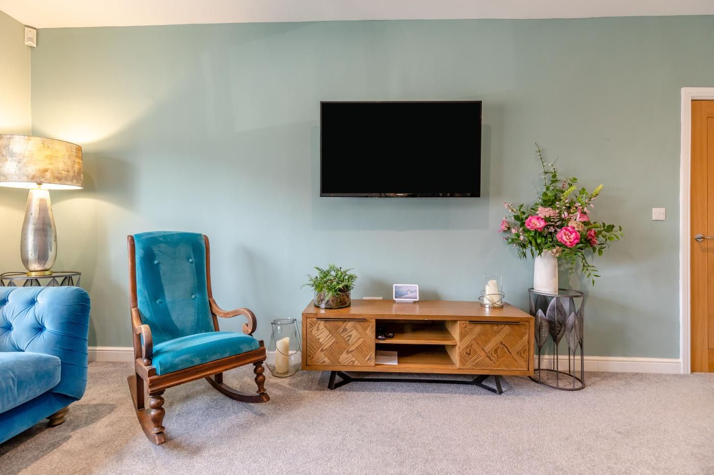 A living room with a blue armchair, a wooden entertainment unit, a flat-screen TV, a lamp, and a vase with flowers.