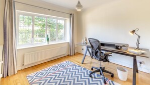 Et værelse med et stort vindue, et skrivebord med stol, en lampe og et mønstret tæppe.
