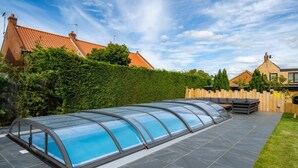 En overdækket swimmingpool med et glasafsnit, omgivet af en terrasse med siddepladser og et træhegn.