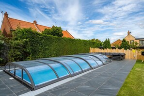 Ein überdachter Swimmingpool mit Glaskuppel, umgeben von einer Terrasse mit Sitzgelegenheiten und einem Holzzaun.