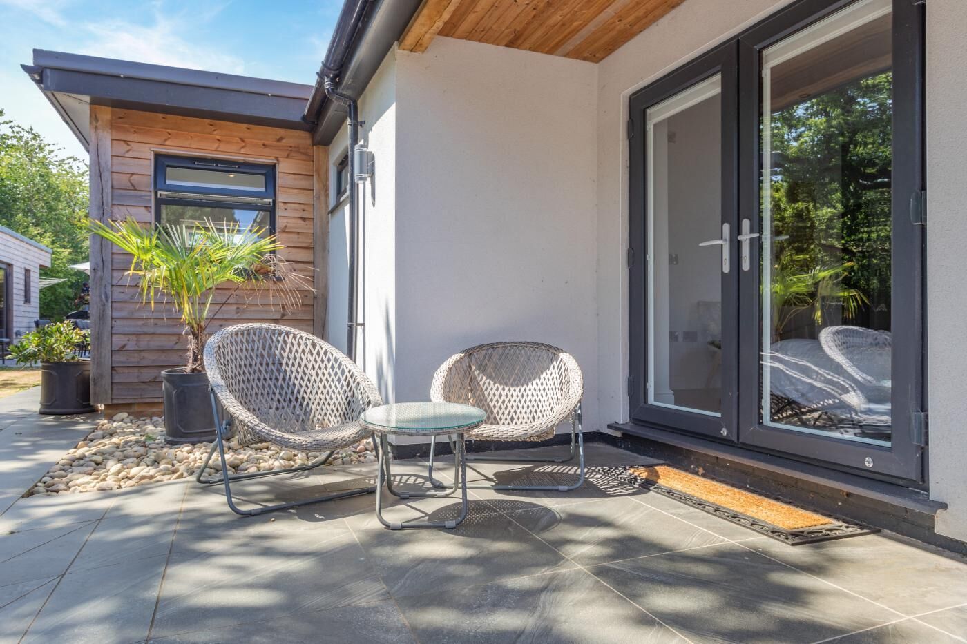 Un espace extérieur moderne, aménagé avec deux fauteuils en osier et une petite table, un mur en bois et une porte-fenêtre coulissante donnant sur un patio abrité.