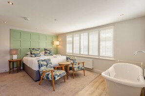 Une chambre d’hôtel comprenant un lit, un coin salon avec des fauteuils, une grande fenêtre avec des stores et une baignoire.