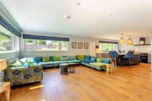 Un vaste espace de vie comprenant un grand canapé en forme de L, une table à manger avec des chaises et un coin cuisine doté de luminaires suspendus.