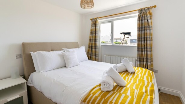 A neatly made bed with white linens and a yellow-striped blanket, a bedside table with a lamp, and a window with curtains. - 3 Bed in Pendeen (oc-g33305) (Pendeen)