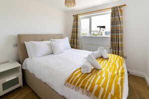 A neatly made bed with white linens and a yellow-striped blanket, a bedside table with a lamp, and a window with curtains. - 3 Bed in Pendeen (oc-g33305) (Pendeen)