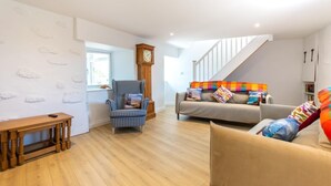 A spacious living room with a wooden floor, a checkered armchair, a sofa with colorful cushions, a wooden bench, and a clock on the wall. - 3 Bed in Pendeen (oc-g33305) (Pendeen)