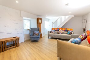 A spacious living room with a wooden floor, a checkered armchair, a sofa with colorful cushions, a wooden bench, and a clock on the wall. - 3 Bed in Pendeen (oc-g33305) (Pendeen)