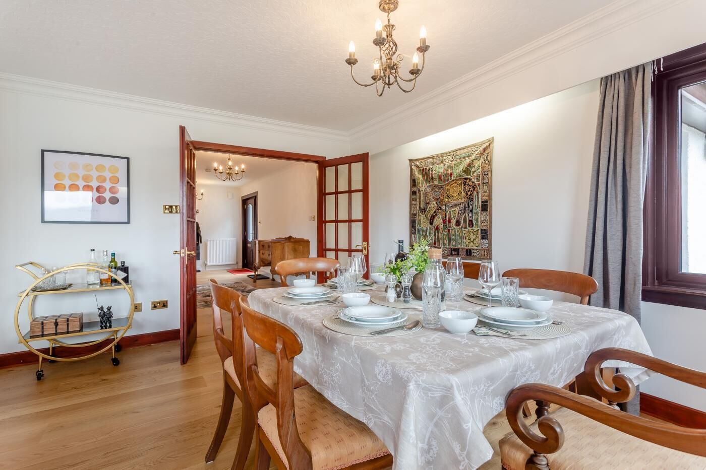 A dining room with a table set for four, wooden chairs, a chandelier, and a decorative tapestry on the wall.