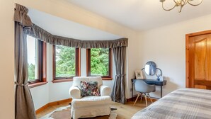 A bedroom with a large window, a white armchair with a floral pillow, a desk with a chair, and a bed with a striped blanket. - 3 Bed in Lairg (oc-r44959) (Little Assynt)