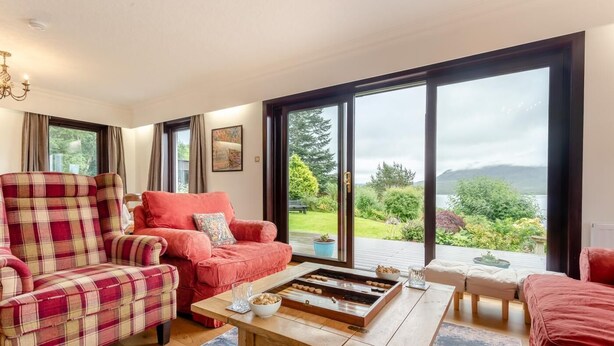 A cozy living room with a plaid sofa, a wooden coffee table, and a view of a lush garden through large windows. - 3 Bed in Lairg (oc-r44959) (Little Assynt)