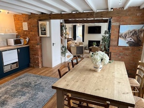 A dining area with a wooden table, chairs, and a vase of flowers. There is a kitchen area with a microwave and a cabinet. A brick wall and a door leading to another room are visible. - 3 Bed in Overton (oc-b45279) (Overton)