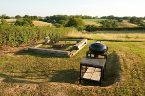 Un'area barbecue all'aperto con tavolo e griglia, immersa in un prato erboso e alberi.