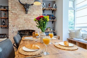 Un comedor con pared de ladrillo, chimenea y mesa dispuesta para el desayuno, con cruasanes, jugo y un jarrón de flores.
