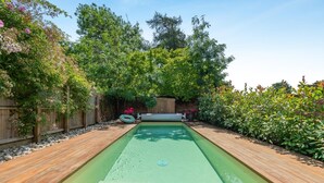 Een rechthoekig zwembad met een houten terras eromheen, gelegen in een tuin met diverse beplanting en een houten omheining.