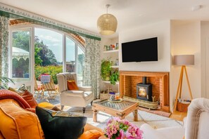 A living room with a fireplace, a television mounted on the wall, and a large window overlooking a garden. - 2 Bed in Great Witchingham (oc-p45227) (Great Witchingham)
