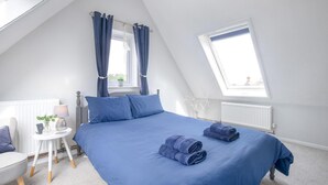A bedroom with a sloped ceiling, a bed with blue bedding, a bedside table with a lamp, and a window with curtains. - 3 Bed in Sheringham (oc-s44832) (Sheringham)