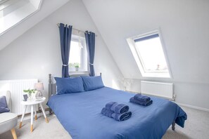 Ein Schlafzimmer mit schrägen Wänden, einem Bett mit blauer Bettwäsche, einem Nachttisch mit Lampe und einem Fenster mit Vorhängen.