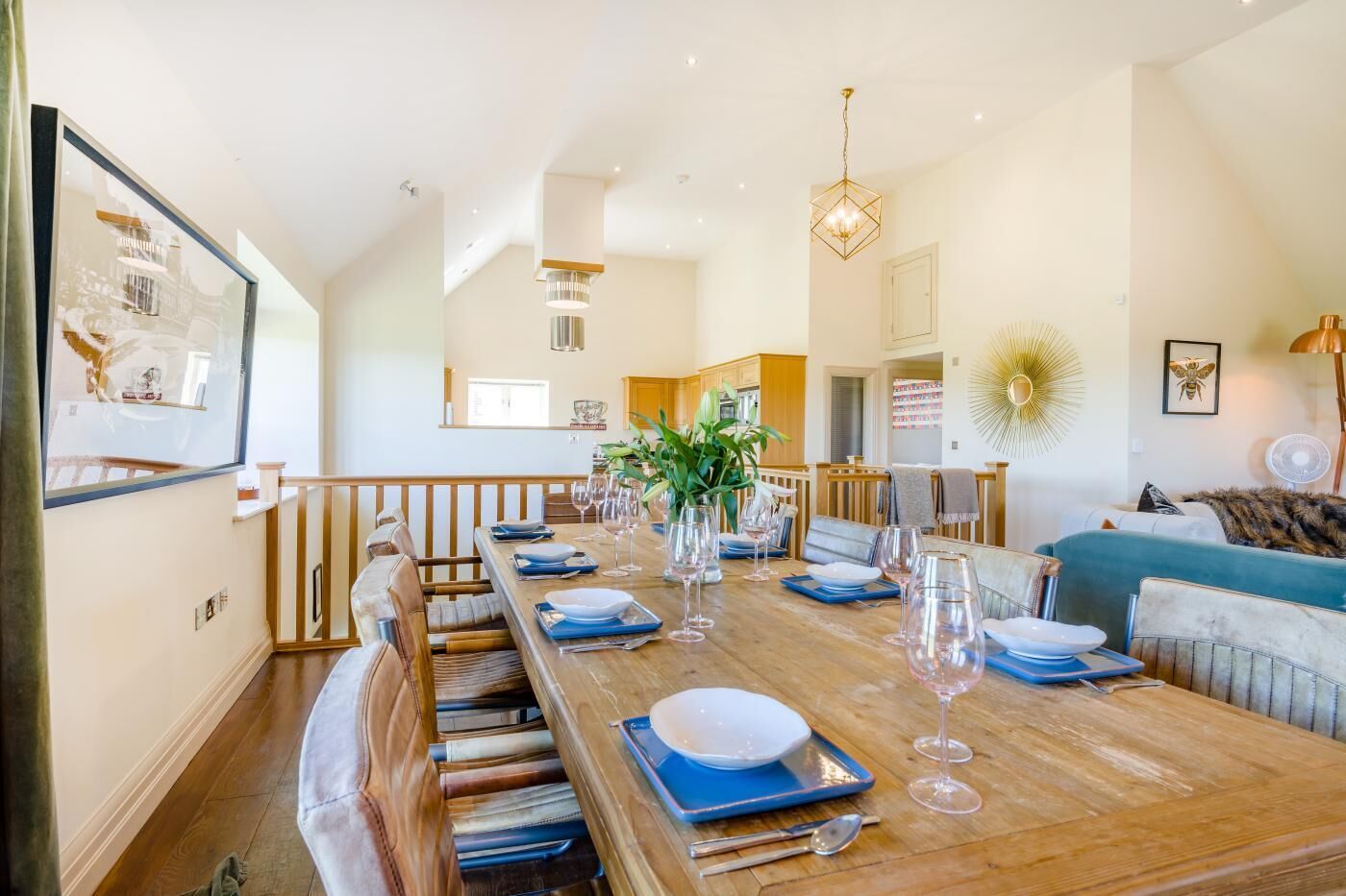 A dining area with a long wooden table set for a meal, a kitchen in the background, and a living area with a sofa and a plant.