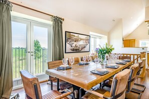 Ein Esszimmer mit einem langen Holztisch, gedeckt für sechs Personen, großen Fenstern mit Vorhängen und Blick ins Grüne.