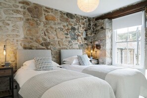 A room with two beds, stone walls, a nightstand with a lamp, and a window with a view. - 2 Bed in Mousehole (oc-b584374) (Mousehole)