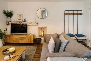 A living room with a wooden TV stand, a sofa, a small table with a candle, and a mirror on the wall. - 2 Bed in Mousehole (oc-b584374) (Mousehole)