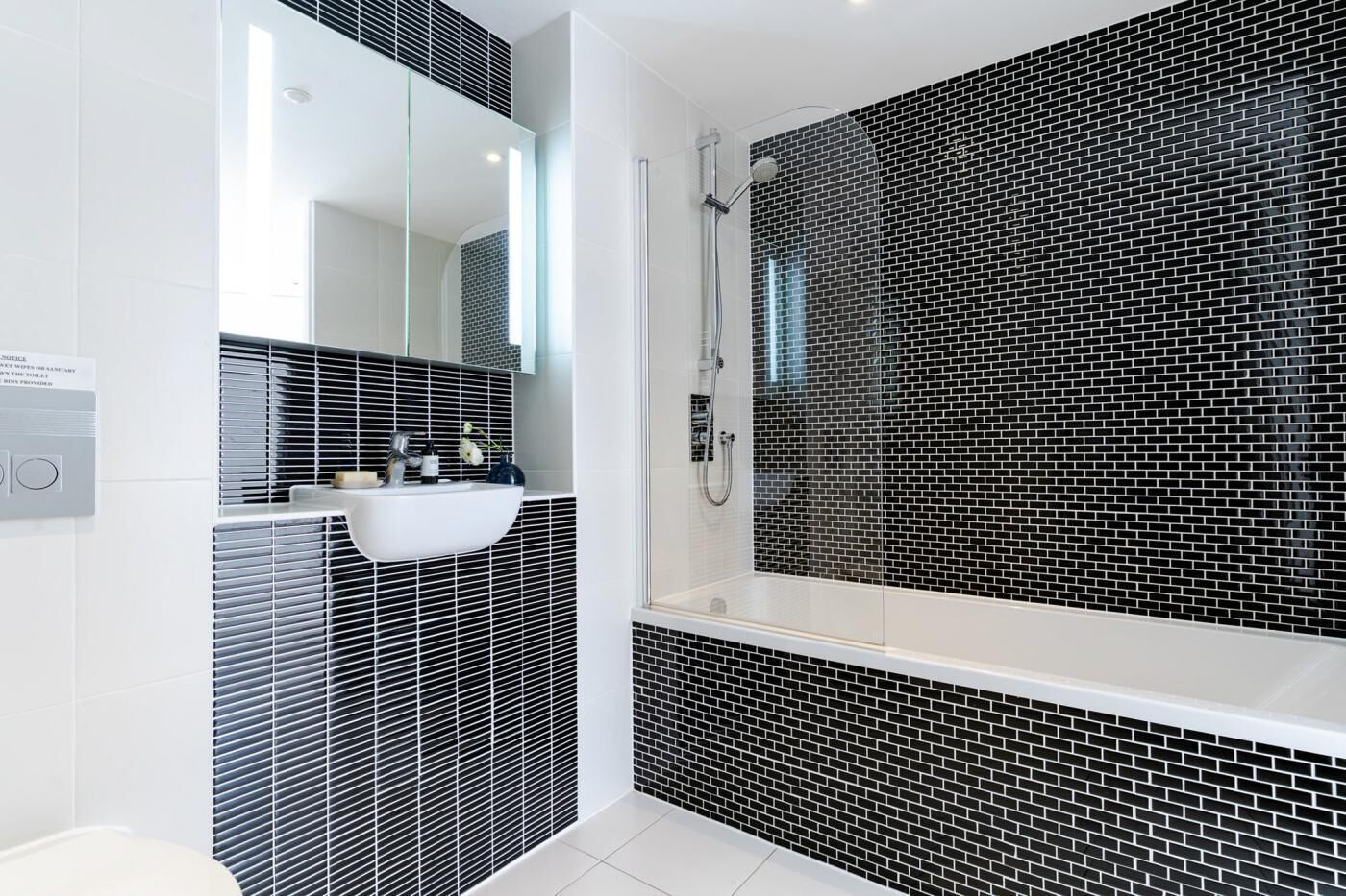 A modern bathroom with a black and white tiled wall, a white sink, and a glass-enclosed shower area.