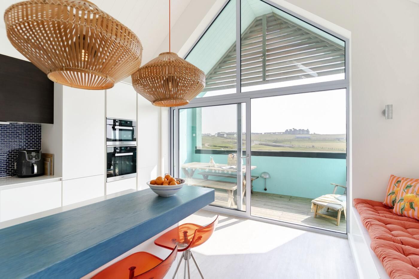 A modern kitchen with a large island, two hanging wicker light fixtures, and a view of a balcony with a table and chairs.