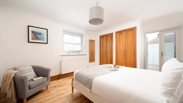 A bedroom with a bed, a grey sofa, wooden wardrobes, a radiator, and a window. - 4 Bed in Porth (oc-b584401) (Newquay)