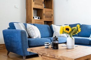 Ein blaues Sofa mit gestreiften Kissen, ein Couchtisch aus Holz mit Kaffeekanne und Tasse sowie eine Vase mit Sonnenblumen.