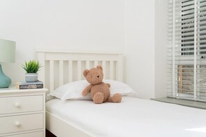Ein weißes Bett mit Teddybär auf dem Kopfkissen, ein Nachttisch mit Lampe und Büchern sowie ein Fenster mit Jalousien.