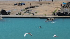 En swimmingpool med krystalklart, blåt vand, omgivet af en sandstrand med sten og mennesker.
