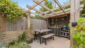 Een houten pergola met een tafel en bankje, omgeven door een tuin met verschillende planten en bloemen.