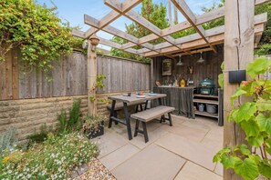 Eine Hollaube mit Tisch und Bank, umgeben von einem Garten mit verschiedenen Pflanzen und Blumen.