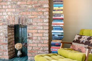 Ein Kamin aus Ziegelsteinen mit einer Vase mit getrockneten Blumen, ein Bücherstapel an einer Wand und ein gelber Sessel mit einem gemusterten Zierkissen.