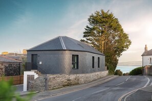 A modern house with a stone foundation and a grey roof, situated on a curved road with a view of the sea. - 2 Bed in Padstow (oc-b623759) (Padstow)