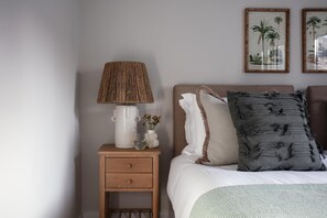 A bedroom with a wooden nightstand, a white lamp, a vase with flowers, a bed with a white and gray pillow, and two framed botanical prints on the wall. - 2 Bed in Padstow (oc-b623759) (Padstow)