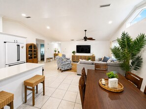 A modern kitchen with white cabinets, a dining area with a wooden table and chairs, a living room with a sofa and armchairs, and a large window with a view. - The Primrose Coastal Retreat •Private Heated Pool • Near Anna Maria Island (Bradenton)