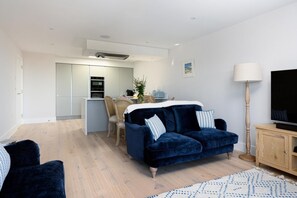 A modern living room with a dark blue sofa, a wooden floor, and a kitchen in the background. - 2 Bed in Porth (oc-b642115) (Newquay)