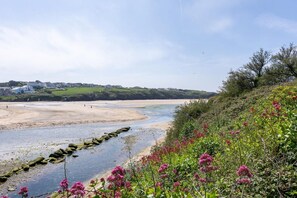 A coastal landscape with a sandy beach, a rocky shoreline, and a hillside with flowering plants. - 2 Bed in Porth (oc-b642115) (Newquay)