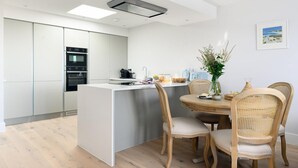 Een moderne keuken met inbouwapparatuur, een eethoek met een ronde tafel en rieten stoelen, en uitzicht op zee vanuit het raam.